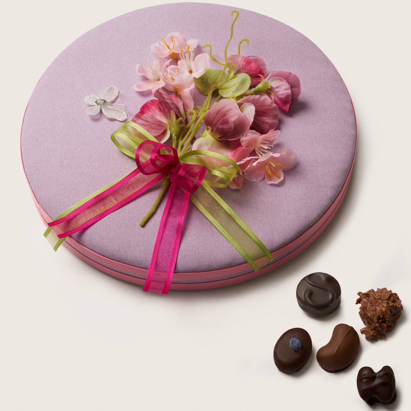 Round pink gift box with floral decoration and ribbons, accompanied by small chocolate truffles on a light background.