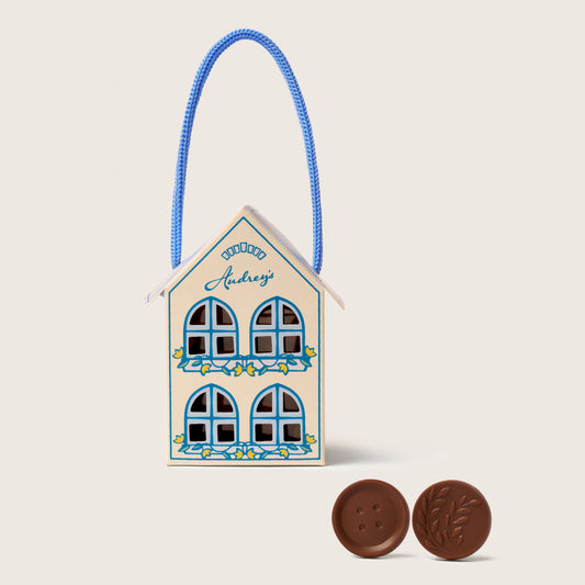 Decorative house-shaped candy container with a blue rope and two chocolate cookies on a beige background