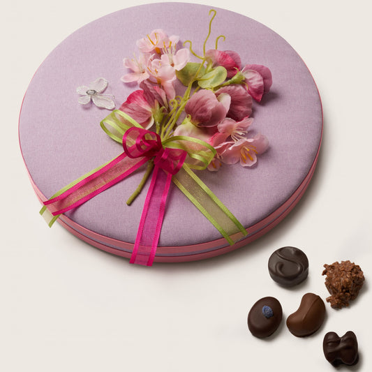 Round pink gift box with floral decoration and ribbons, accompanied by small chocolate truffles on a light background.