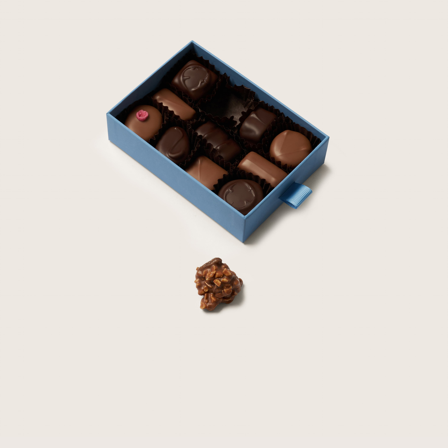 Open blue box of assorted chocolates with a single chocolate on a light gray background